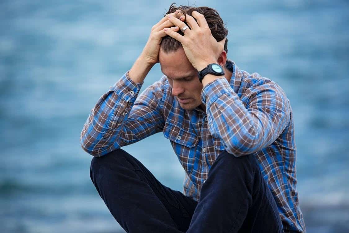 A Man Sitting by a Body of Water and Holding His Head in His Hands in Visible Distress