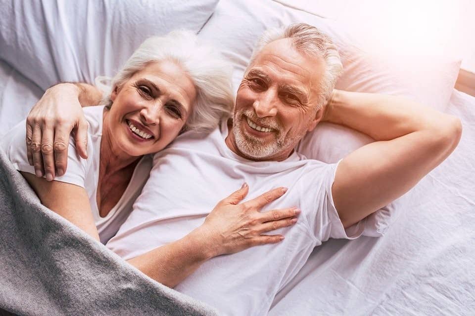 A Visibly-Blissful Senior Couple Cuddling Up in Bed While Staring Right at the Camera