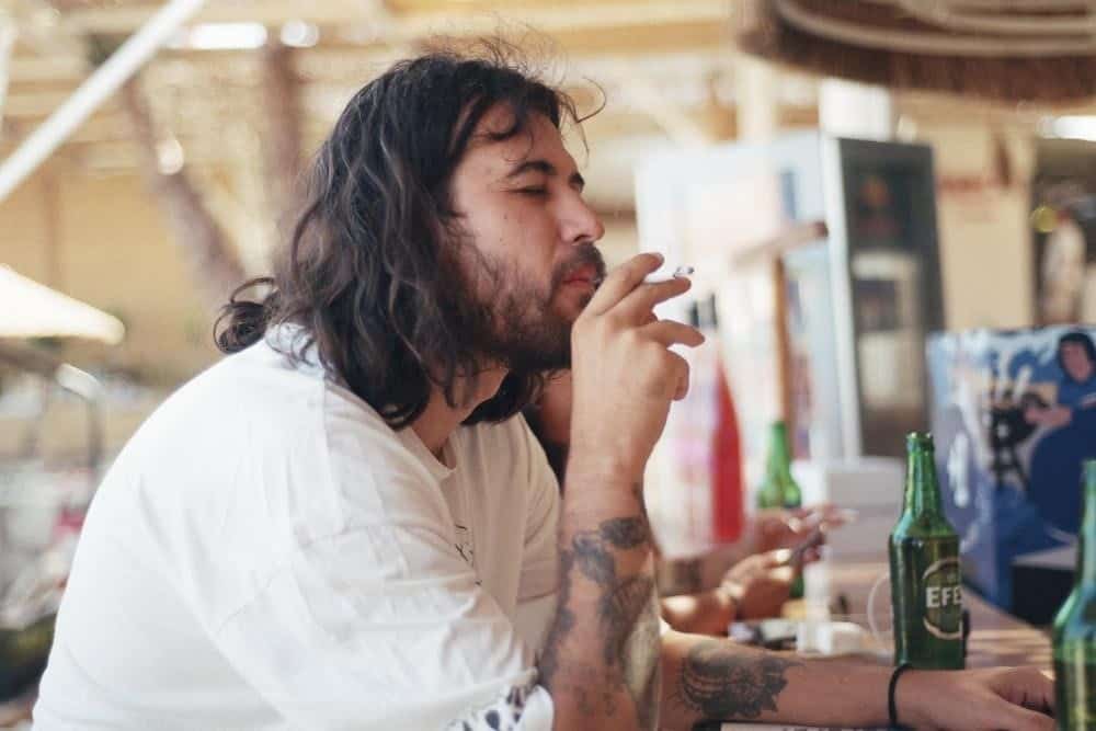 A Man Sat at a Bar and Having a Smoke While Holding a Beer Bottle